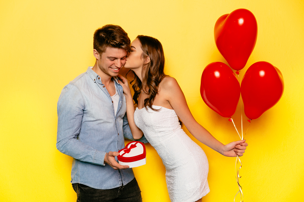 Beautiful girl dressed in white dress awith red balloons kissing her boyfriend holding box with gift inside. St. Valentine's day